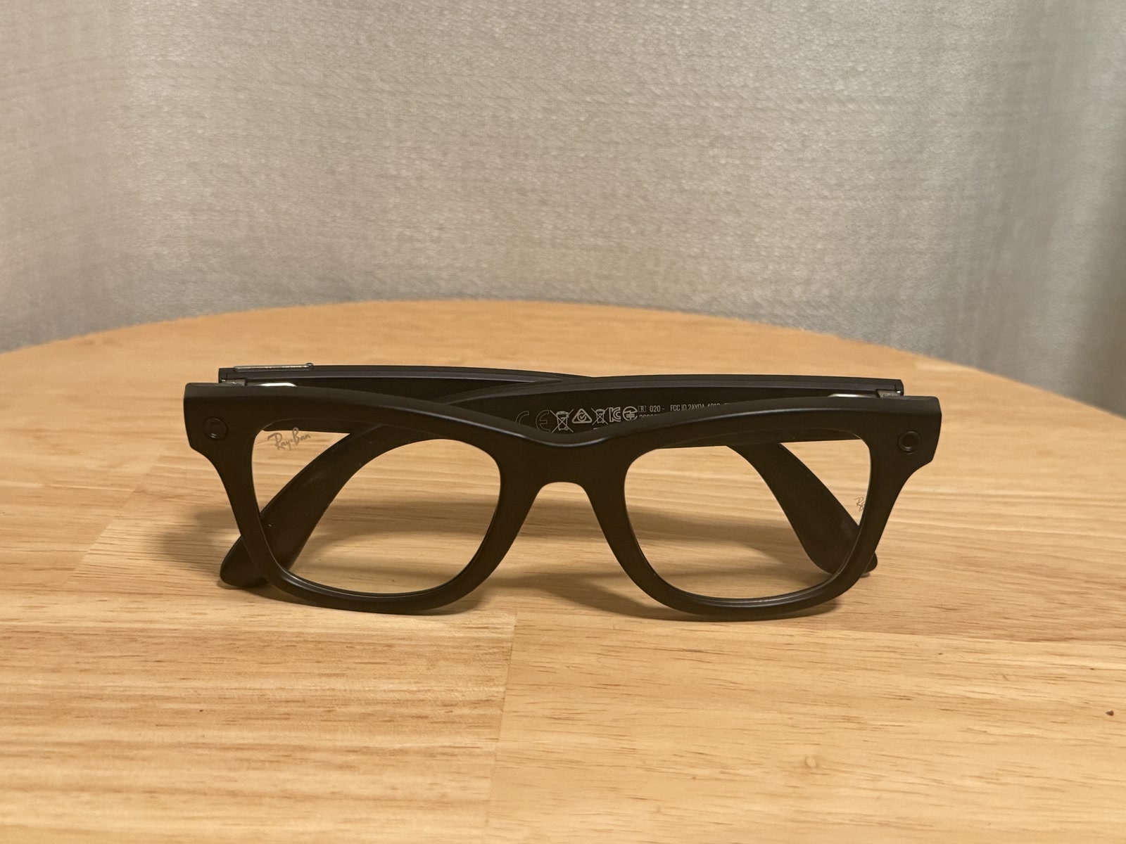 Ray-Ban Meta Gen 3 Wayfarer frontal auf dem Holztisch, Bügel eingeklappt, kleine Kamerapunkte an beiden Ecken